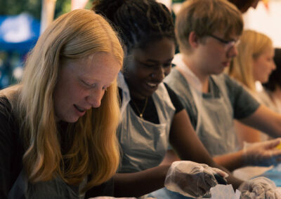 Een groep jongeren doet enthousiast mee aan een tie-dye workshop op een zomerkamp. Ze dragen schorten en handschoenen terwijl ze witte shirts met kleurrijke verf bewerken. Op de achtergrond hangen feestelijke vlaggetjes onder een tent.